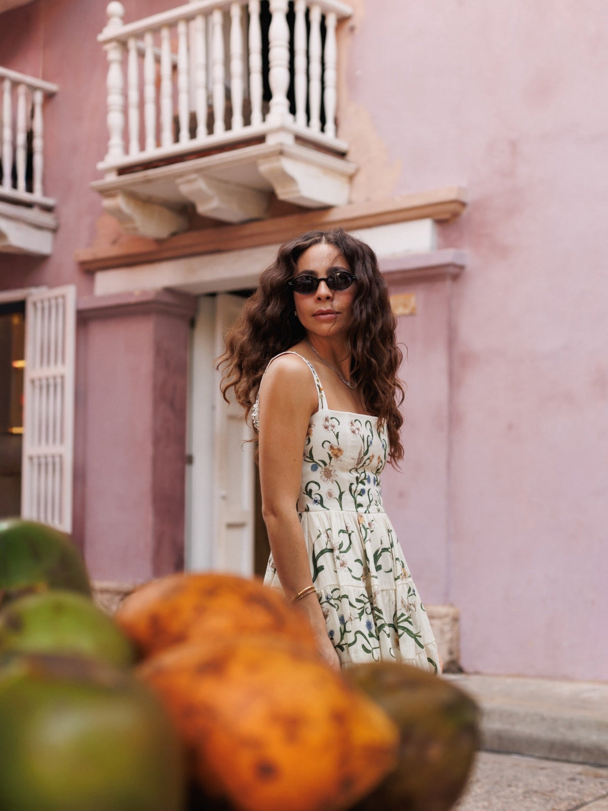 Woman in sunglasses and a white botanical-print sundress outside a pink stucco building